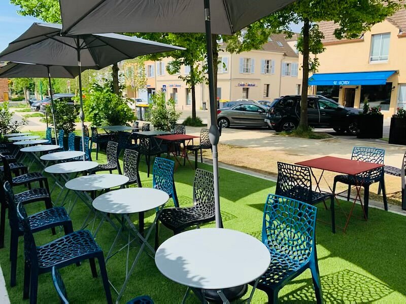 Terrasse extérieure du Gondi : tables blanches, chaises bleues et parasols sur gazon, devant des bâtiments anciens.